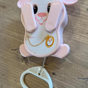 May include: A pink and white wind-up musical toy shaped like a rabbit with a pull string and a clock face on its chest. The rabbit is holding its paws up to its face.