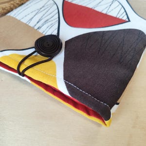 Può includere: Una custodia in tessuto con chiusura a bottone. La custodia presenta un design colorato con forme marroni, rosse, gialle e bianche. Un bottone e un elastico marrone scuro fissano la custodia. La fodera interna è rossa.