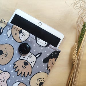 Può includere: Una custodia grigia per tablet con un motivo di facce di cani dei cartoni animati beige e marroni. La custodia ha una chiusura con bottone nero e contiene un tablet bianco. Lo sfondo è una superficie di legno chiaro.