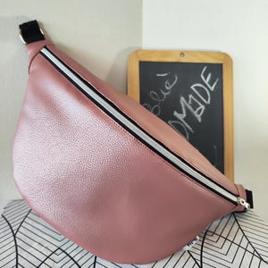 Può includere: Un marsupio rosa cipria con finitura strutturata. La borsa presenta una cerniera nera con una striscia bianca e un cinturino nero. La parola "MADE" è scritta su una piccola lavagna sullo sfondo.