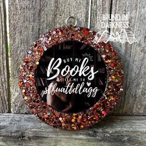 May include: A round, copper-colored glitter ornament with a black and white photo inside. The photo shows a bookshelf with books and a person's hand reaching for a book. The text on the ornament reads "Buy me books & tell me to shut up and read."