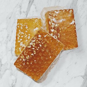 Puede incluir: Tres bloques rectangulares de material ámbar texturizado, posiblemente jabón o cera, están envueltos en plástico transparente. Los bloques tienen un aspecto moteado y el embalaje es transparente.