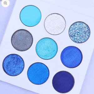 May include: A square eyeshadow palette featuring nine circular pans arranged in a grid. The colors include shades of sky blue, silver, dark gray, metallic blue, deep navy, and a glittery blue texture, all set within a plain white casing.