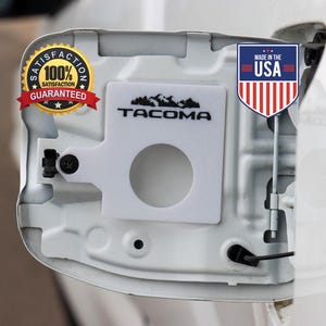 May include: White car part with the word "TACOMA" in black lettering, a mountain graphic, and a circular opening. Includes a "Made in the USA" emblem and a "100% Satisfaction Guaranteed" sticker.