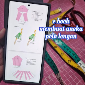 Puede incluir: Un libro electrónico titulado "membuat aneka pola lengan" se muestra en una tableta, mostrando ilustraciones de patrones de mangas. También se ven unas tijeras y varias cintas métricas amarillas y verdes, lo que sugiere un tema de costura o diseño.