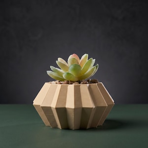 May include: A light beige, geometric, faceted planter with a wide, multi-sided base. Inside the planter sits a small, artificial succulent plant with thick, fleshy, light green and pink-tipped leaves, surrounded by small, smooth brown pebbles.