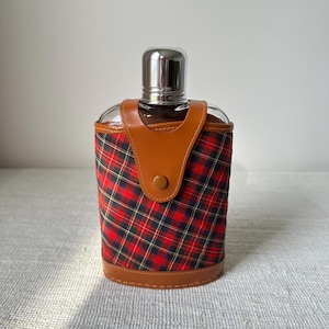 May include: A glass flask with a stainless steel cap, encased in a red, black, and yellow plaid fabric and brown leather accents. The flask has a brown leather flap with a button closure.