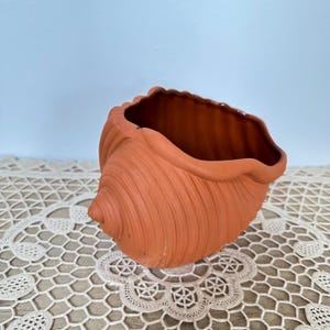 May include: A terracotta-colored seashell-shaped planter. The planter has a textured, ridged exterior and a smooth, brown interior. The rim of the planter has a scalloped edge. The planter is sitting on a cream-colored, lace doily.