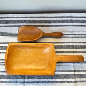 May include: Two wooden kitchen utensils: a scoop and a rectangular tray with a handle. Both are made of light-colored wood and rest on a striped fabric. The scoop has a rounded bowl and a short handle. The tray has a rectangular shape with rounded corners.