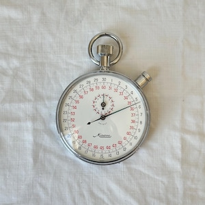May include: A vintage stopwatch with a white face, black and red numerals, and a silver metal casing. The stopwatch has a crown at the top and a button on the side. The brand name "Minerva" is printed on the face.