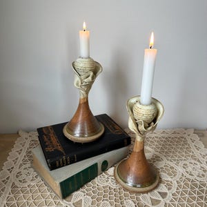 May include: Two ceramic candle holders with lit white candles. The candle holders have a unique design with a brown and beige color scheme. They are placed on top of two vintage books.