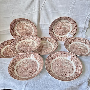 May include: A set of seven round ceramic bowls featuring a brown transferware pattern. Each bowl displays a detailed landscape scene with a cathedral, trees, and figures, surrounded by a decorative floral border on the rim and interior.