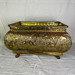 May include: A rectangular brass planter featuring an embossed floral pattern on the sides and a decorative cutout border along the top rim. The vessel is supported by four small, curved feet and has a polished, reflective metallic finish.