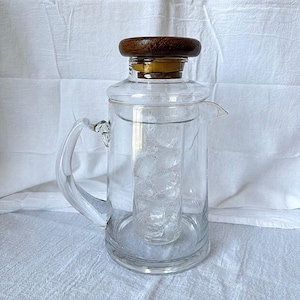 May include: A clear glass pitcher featuring a central cylindrical ice chamber filled with ice cubes. The pitcher has a curved glass handle, a pouring spout, and a circular dark brown wooden lid with a visible grain, secured by a yellow seal.