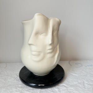 May include: A cream-colored ceramic vase featuring three sculpted faces. The vase is set on a round, black base. The faces are smooth and detailed, with subtle shadows. The overall design is artistic and modern, suitable for home decor.