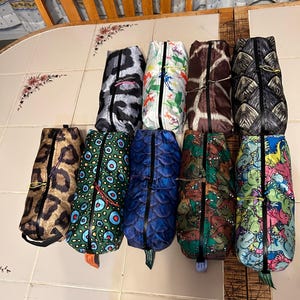 May include: A collection of ten zippered pouches with various patterns, including animal prints like leopard and giraffe, and designs featuring dinosaurs and abstract shapes. Each pouch has a black zipper and a small loop at the bottom.