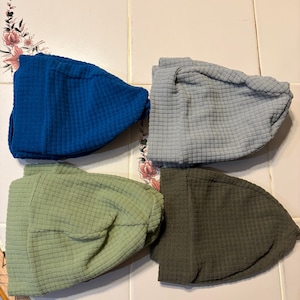May include: Four textured fabric hats in shades of blue, gray, green, and olive. The hats have a quilted pattern and are arranged on a tiled surface. The hats appear to be a winter accessory.