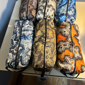 Peut inclure: Six pochettes zippées à motifs différents avec des tirettes de fermeture éclair noires. Les pochettes sont fabriquées dans un tissu imperméable et présentent une variété de couleurs et de motifs, notamment le bleu, le vert, l'orange et le marron.