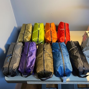 May include: A collection of nine rectangular zippered pouches in various colors including olive, lime green, orange, red, gray, purple, brown, blue, and black. Each pouch features a central black zipper, a pull tab, and a structured boxy shape.