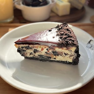 May include: A single slice of cookies and cream cheesecake on a white plate. The cheesecake features a dark chocolate cookie crust, a creamy filling marbled with dark cookie pieces, and a smooth dark chocolate ganache topping with cookie crumbs.