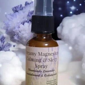 Pode incluir: Um frasco de spray de vidro castanho com uma tampa preta e topo transparente. O rótulo diz "Dreamy Magnesium Calming & Sleep Spray" com os ingredientes listados. O fundo apresenta um céu noturno estrelado e nuvens fofas.