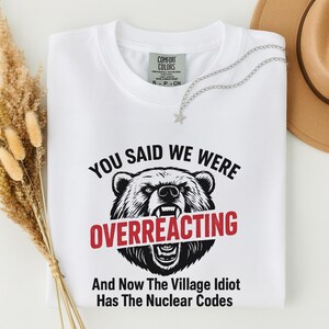 Pode incluir: Camiseta branca com um gráfico de cabeça de urso e o texto "YOU SAID WE WERE OVERREACTING" em preto e vermelho. Abaixo do gráfico do urso, o texto diz "And Now The Village Idiot Has The Nuclear Codes". Um colar de prata com um pingente de estrela está na camisa.