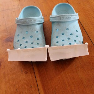 Peut inclure: Une paire de Crocs bleu clair avec des attaches en plastique blanc sur le dessous. Les Crocs ont le mot "Crocs" imprimé sur la sangle.
