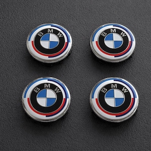 Op de afbeelding: Vier ronde BMW-emblemen met een zilveren rand. Elk embleem toont het BMW-logo, een blauw-wit geblokt patroon omcirkeld door zwarte, rode en blauwe ringen, en de letters "BMW".