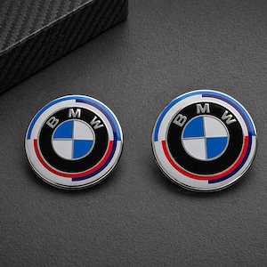 Könnte beinhalten: Zwei kreisförmige BMW-Embleme. Jedes Emblem hat eine blau-weiß karierte Mitte, umgeben von einem schwarzen Ring, einem rot-blauen Streifen und einem blauen Außenring. Die Buchstaben "BMW" sind abgebildet.