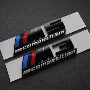 Pode incluir: Dois emblemas M3 Competition pretos e prateados com riscas azuis, vermelhas e prateadas. Os emblemas estão em tiras de suporte brancas, sobre um fundo cinzento escuro.