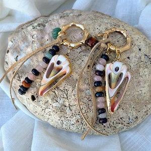 May include: A pair of seashell earrings with gold-colored hoops and tan leather cords. Each earring features a shell pendant with a colorful interior and is adorned with a strand of beads in various colors, including pink, black, and brown.