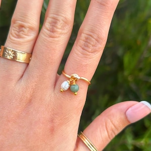 May include: A gold-colored ring with a small cluster of white and green beads. A second gold ring with a starburst design is also visible. The rings are on a hand with a light pink manicure.