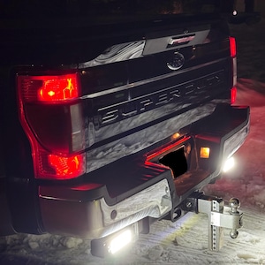 May include: Rear view of a black pickup truck with a chrome bumper and a hitch receiver. The truck has red taillights and a "SUPER DUTY" emblem on the tailgate. The hitch receiver has a ball and a safety chain.