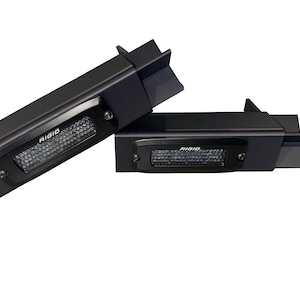 Può includere: Due apparecchi di illuminazione a LED rettangolari neri con la scritta "RIGID" stampata sulla parte anteriore. Gli apparecchi hanno un motivo strutturato a griglia al centro e sono progettati con bordi angolati.