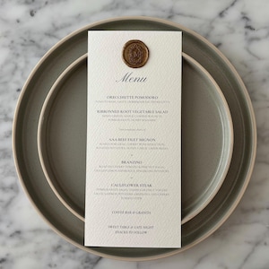 May include: A formal menu card with a gold wax seal, placed on a gray plate. The menu lists dishes like Orecchiette Pomodoro and Cauliflower Steak. The card is centered on a larger plate, set against a marble background.