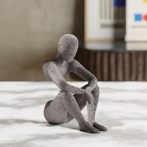 May include: A gray, abstract sculpture of a person sitting with their legs crossed. The sculpture has a rough, textured surface.