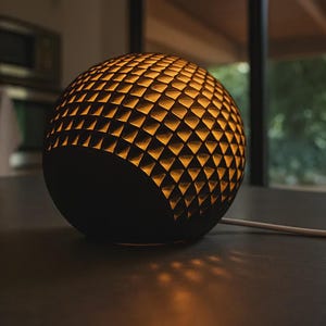 May include: A black spherical lamp with a geometric diamond pattern. The lamp emits a warm, golden light through the cut-out design. A white power cord extends from the base. The lamp is on a dark surface.
