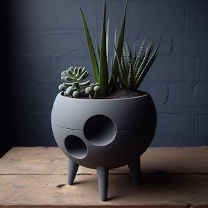 May include: A gray, spherical planter with three legs and three circular cutouts. The planter holds various succulents and aloe plants with green leaves. The planter sits on a wooden surface against a dark blue brick wall.