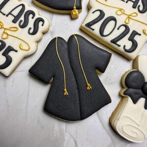 May include: Graduation-themed cookies. A black graduation gown cookie with gold accents is surrounded by other cookies with the text "CLASS 2025" and "2025" in black icing. The cookies are on a white surface.