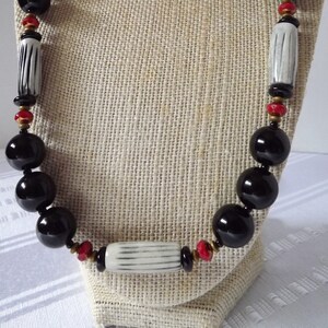 May include: A black and white beaded necklace with red accents. The necklace is made of round black beads, white and black striped cylinder beads, and small red beads. The necklace is displayed on a fabric-covered mannequin.