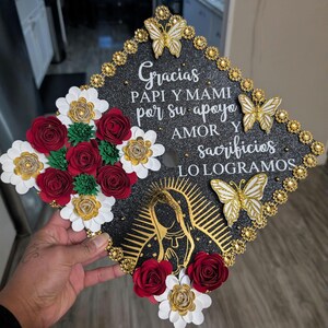 Puede incluir: Un gorro de graduación decorado con un fondo negro brillante. Presenta flores de papel en rojo, blanco y dorado, junto con mariposas doradas y un diseño dorado de la Virgen María. El gorro tiene texto en blanco.