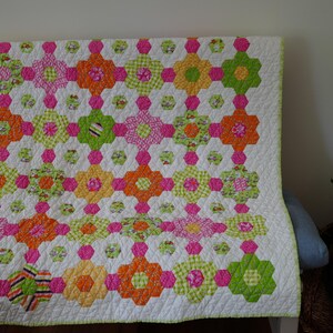 May include: A handmade quilt featuring a repeating pattern of hexagonal flower shapes in vibrant shades of orange, lime green, and hot pink. The quilt has a white background with a subtle quilted texture and a light green border trim.