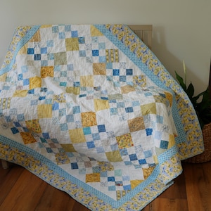 May include: A patchwork quilt featuring a repeating pattern of white, yellow, and blue fabric squares. The quilt is bordered by a wide band of yellow and blue floral print fabric, with smaller blue and white patterned squares throughout the design.
