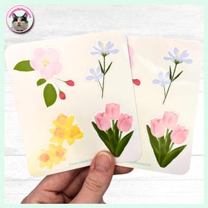 May include: Two sheets of floral stickers featuring various flower designs. The stickers include pink tulips, yellow daffodils, light blue flowers, and a pink flower with green leaves. The stickers are held by a hand.