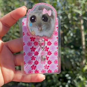 May include: A pink and white floral patterned badge holder with a clear plastic front. The holder features a small, round, clear plastic charm with a cartoon hamster wearing a pink bow and holding a cup of iced coffee. The hamster has a sad expression and a single tear on its cheek.