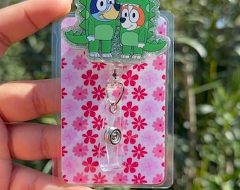 May include: A pink and white floral patterned badge holder with a clear plastic cover. The holder has a retractable badge reel with a silver clip. The top of the holder features a green glitter dinosaur with two cartoon characters inside.