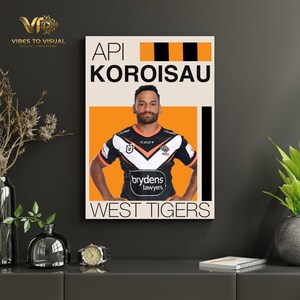 Op de afbeelding: Een ingelijste poster met Api Koroisau in een West Tigers-shirt. De poster heeft een oranje en zwart ontwerp met de naam "API KOROISAU" bovenaan en "WEST TIGERS" onderaan. Het shirt toont het "brydens lawyers" logo.