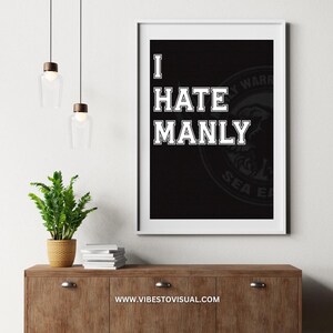 May include: A framed black and white print with the text "I HATE MANLY" in bold, white, block letters. The print is displayed on a wooden cabinet, with a small potted plant and decorative lighting above. The website address "www.vibestovisual.com" is at the bottom.