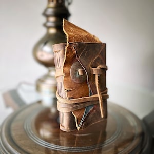 May include: A small, brown leather-bound journal with a worn, rustic look. The journal is closed and tied with a leather strap. The journal is sitting on a wooden surface.