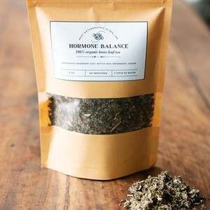 Könnte beinhalten: Ein brauner Papierbeutel mit einem Etikett, auf dem steht "ROOT NATUROPATHIC CLINIC, INC. HORMONE BALANCE 100% organic loose leaf tea INGREDIENTS: RASPBERRY LEAF, NETTLE LEAF, PEPPERMINT, GINGER 3 OZ 40 SERVINGS 2 TSP/B OZ WATER". Der Beutel ist mit losen Blättern gefüllt und steht auf einer Holzoberfläche.